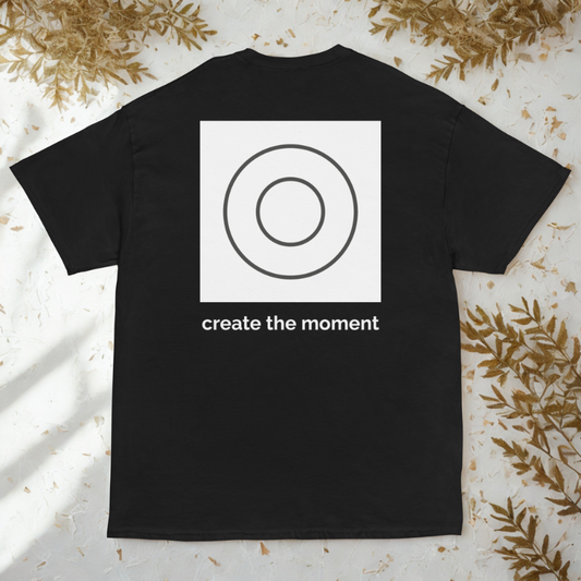 Back view of charcoal tee with white square and grey double-circle logo above “create the moment,” leaf-strewn light terrazzo.