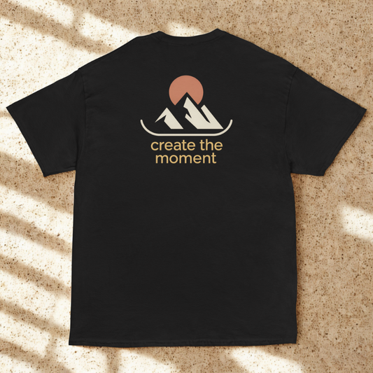 Back view of charcoal tee with minimalist twin peaks and sun icon and “create the moment” slogan, soft dappled window shadows on terrazzo.