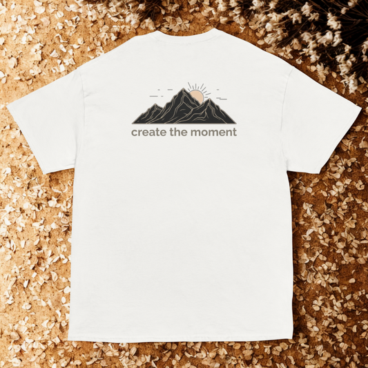 Back view of white tee with line-art mountains and rising sun plus “create the moment” text, warm sand surface with tiny shells/seed pods.