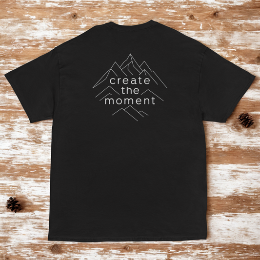 Back view of charcoal tee with thin outline mountain stack framing “create the moment,” rustic wood backdrop with a few pinecones.