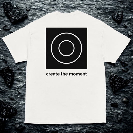 Back view of white tee with black square and double-circle logo above “create the moment,” dark river rock backdrop.