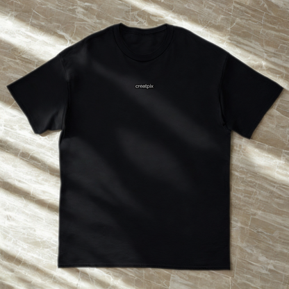 Front view of black classic tee with small white creatpix chest logo, soft window-light on stone surface.