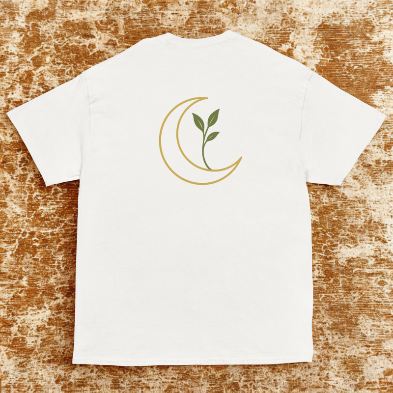 Back view of white tee with gold crescent moon and green sprouting leaves, warm distressed rug backdrop.