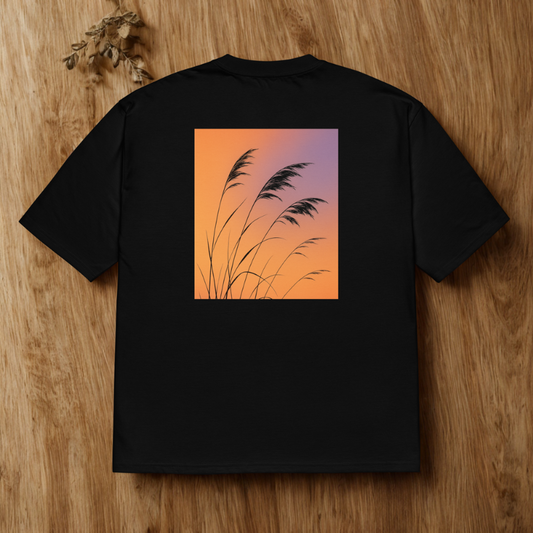 Back view of charcoal tee with square gradient sky and tall grass silhouettes, warm wood surface.