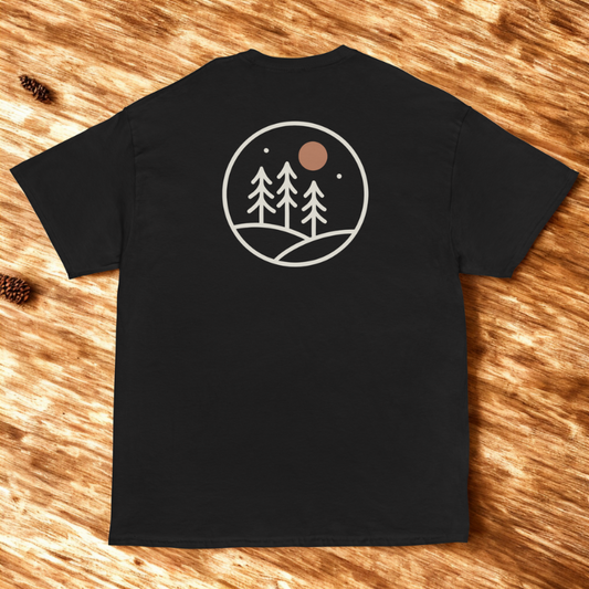 Back view of charcoal tee with pine tree silhouette and retro sunset circle graphic, soft shadows on neutral stone.