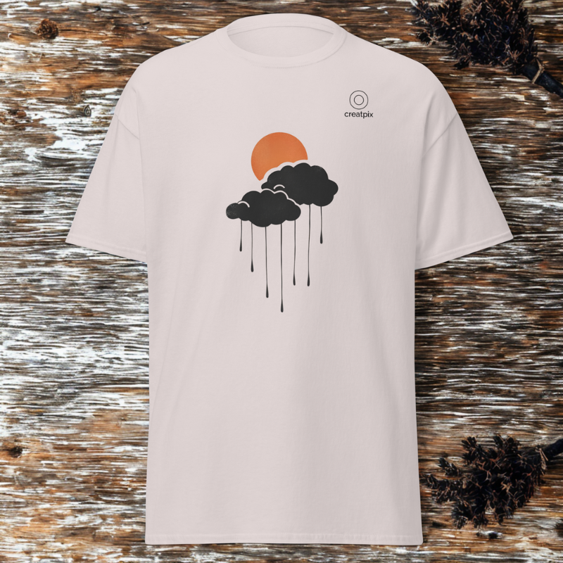 Front view of sand tee with orange sun and dark rain cloud graphic with falling lines, small creatpix chest mark, rustic wood backdrop with pinecones.