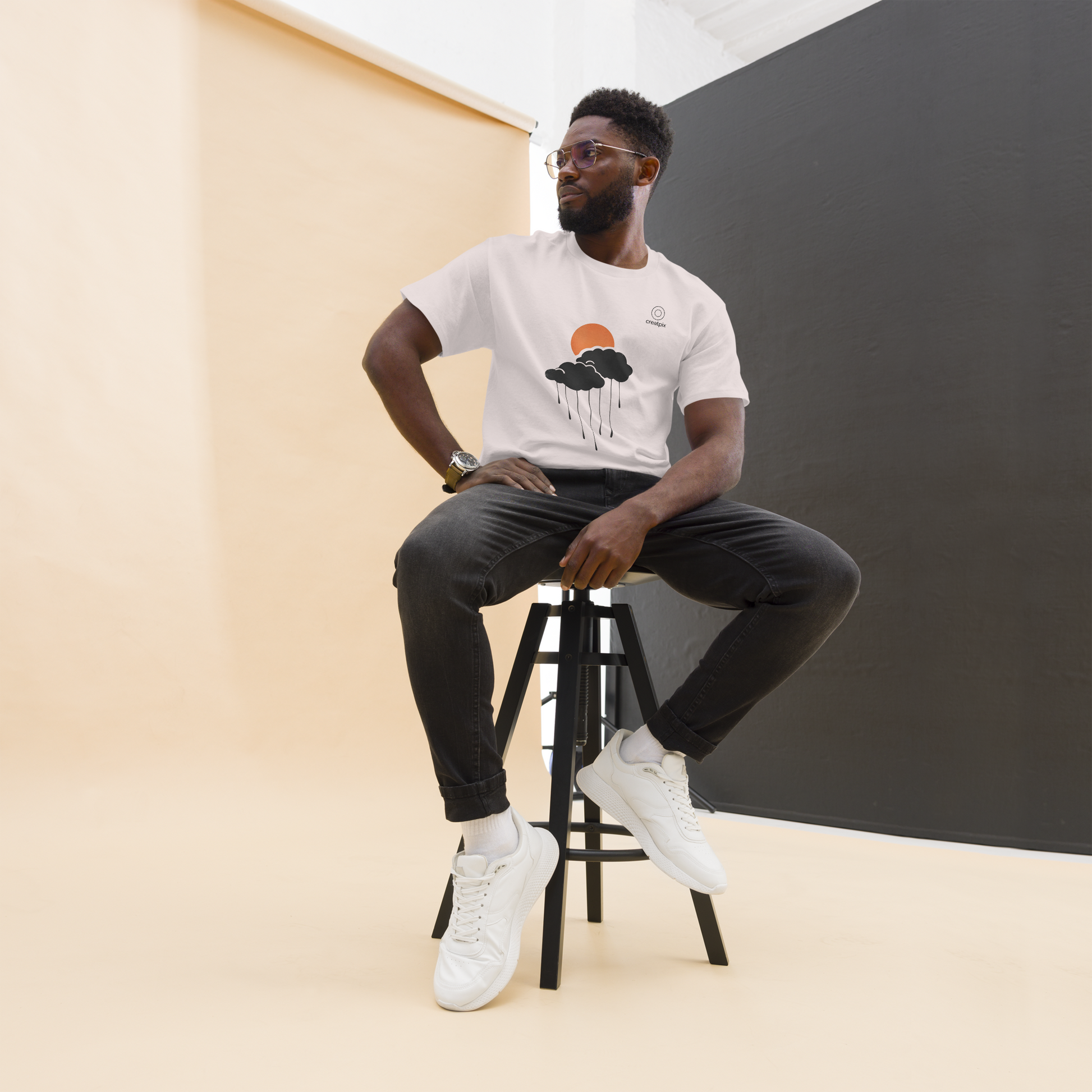 Lifestyle studio shot of model seated on a stool wearing the sand sun-and-cloud rain tee, minimal warm setup.