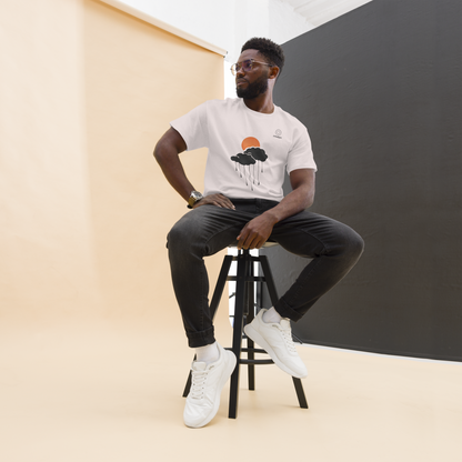 Lifestyle studio shot of model seated on a stool wearing the sand sun-and-cloud rain tee, minimal warm setup.