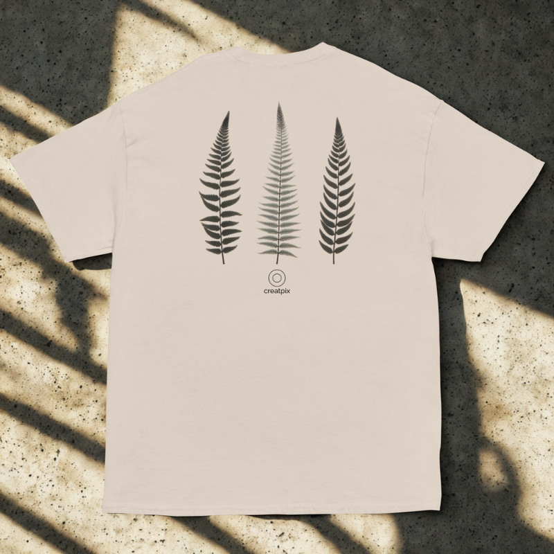 Back view of sand tee with three vertical fern fronds and small round logo, soft window shadows on speckled concrete.