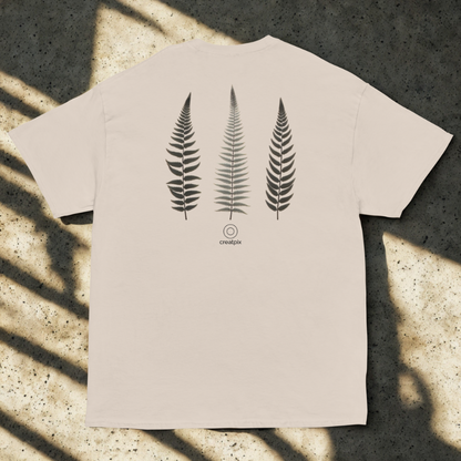 Back view of sand tee with three vertical fern fronds and small round logo, soft window shadows on speckled concrete.