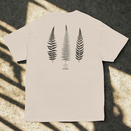 Back view of sand tee with three vertical fern fronds and small round logo, soft window shadows on speckled concrete.